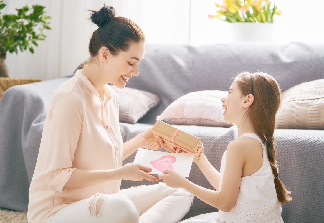 A young girl giving a woman a cark with pink heart and present wrapped in brown paper